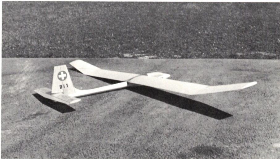 A black and white photograph of the RC glider DIONYS on a flat surface. The glider is shown from a side profile, highlighting its long wings and tail section. The tail features a cross symbol and the number '011'.