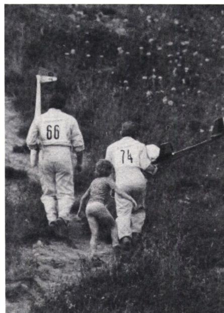 Two people in white flight suits with numbers 66 and 74 on their backs are walking away from the camera on a grassy hillside. They appear to be carrying or preparing to launch model airplanes.