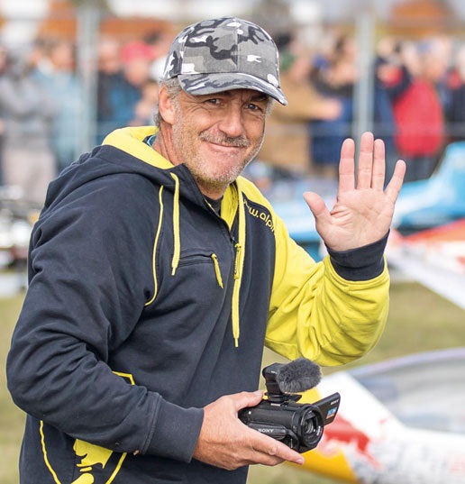 Markus Nussbaumer, wearing a camouflage cap and a yellow and black jacket, holding a Sony Handycam and waving with his other hand at an outdoor event.
