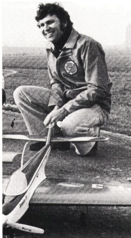 A man kneeling on the ground, holding a model airplane, smiling at the camera.