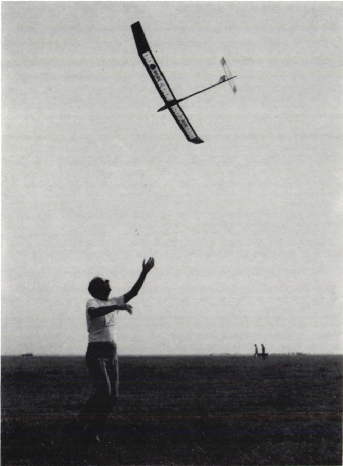 A black and white photograph of a person standing in a field, looking up at a glider plane in the sky. The person's arms are raised in a gesture of greeting or acknowledgment.