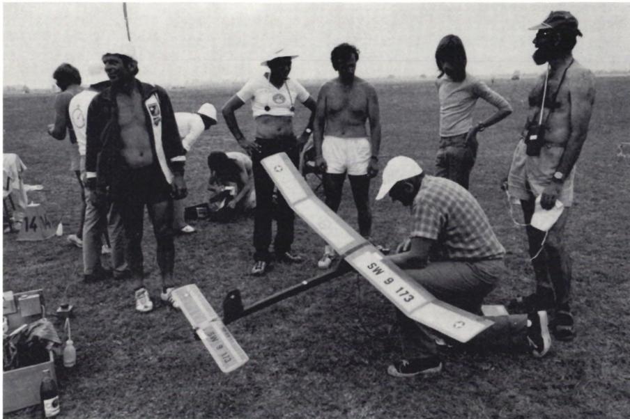 A group of people, including some shirtless men, gathered around a small aircraft on a grassy field. One man is kneeling and working on the aircraft, while others stand nearby.