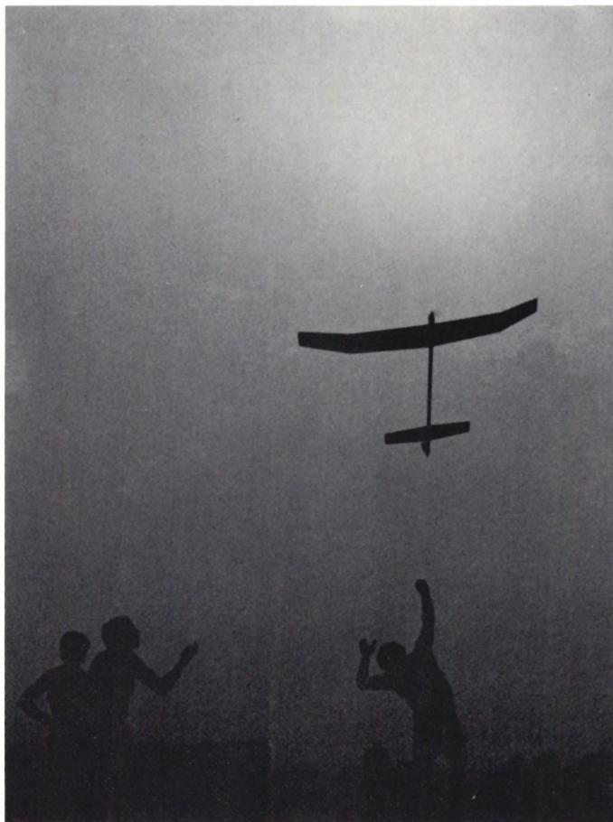 Black and white photograph of a model airplane in flight against a bright, hazy sky. In the foreground, the silhouettes of several people are visible, some with arms raised in excitement.