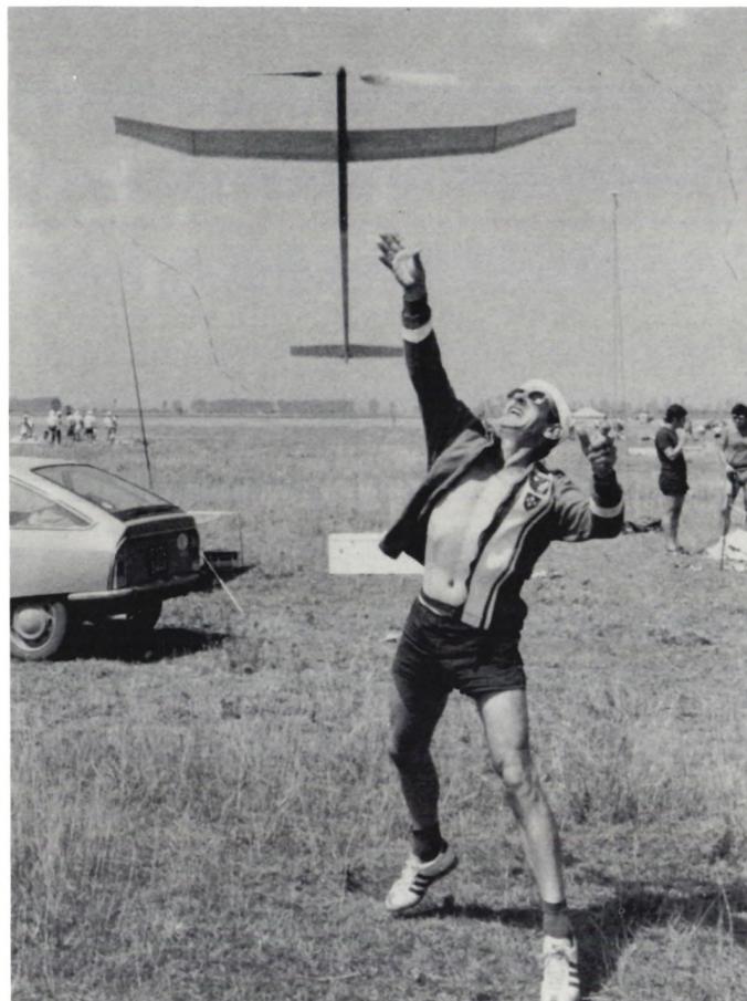 Black and white photograph of Fritz Gänsli, a man in a jacket and shorts, throwing a rubber-powered model airplane into the air. The plane is in flight above him. A car is visible in the background on the left.
