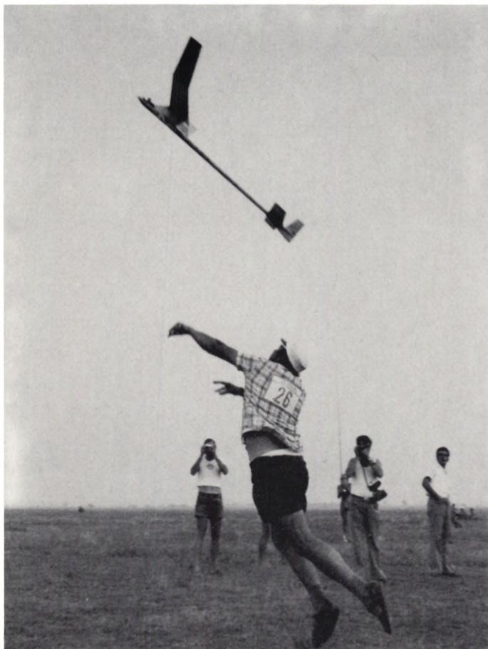 Black and white photograph of a man in a plaid shirt and shorts throwing a model airplane into the air. He is wearing a bib with the number 26. Several other people are standing in the background on a flat, open field.