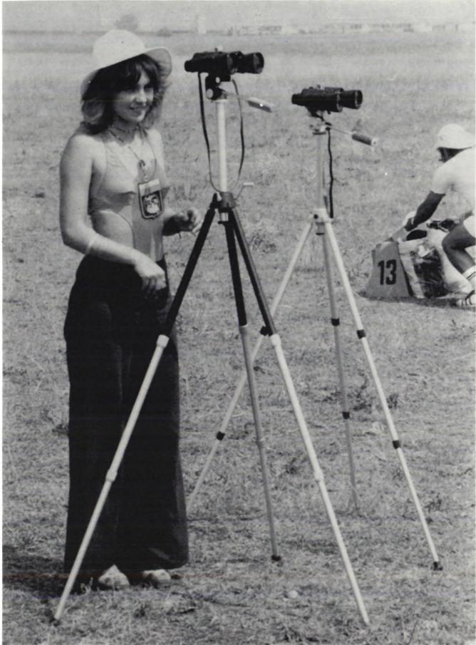 A black and white photograph of a woman wearing a hat and a dark dress, standing next to two tripods with binoculars or cameras. She is smiling at the camera. In the background, another person is visible near a marker with the number 13.
