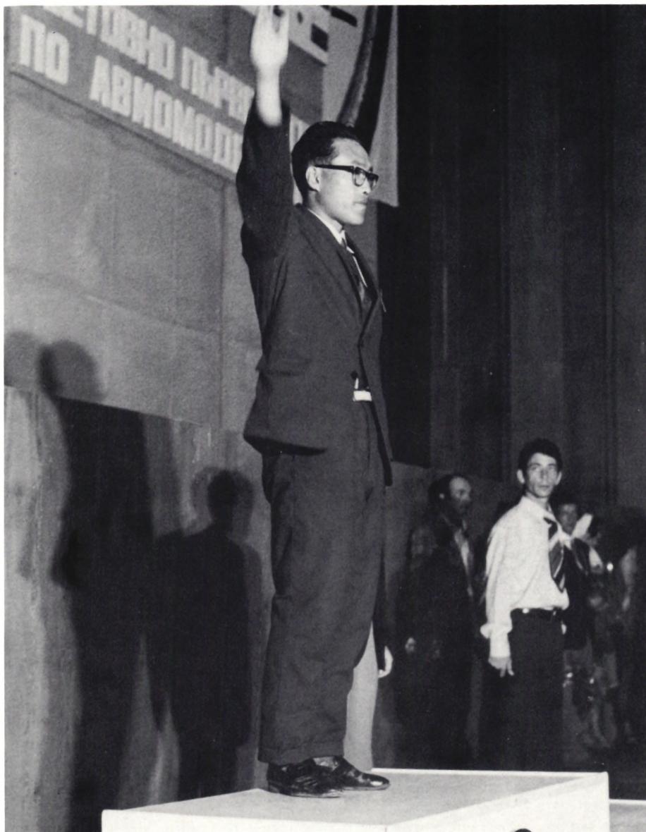 Black and white photograph of Paik Chang Sun, a North Korean射擊手, standing on a podium during the award ceremony. He is wearing a dark suit and glasses, and is raising his right hand in a gesture of acknowledgment. Behind him, a banner with Cyrillic text is visible. Other people are standing in the background, some looking towards the camera and others away.