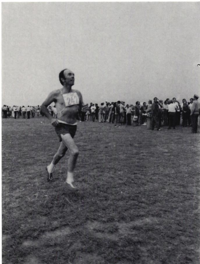 A black and white photograph of a man, Peter Allnutt, running across a grassy field. He is wearing a white athletic singlet with the number '247' and dark shorts. A large crowd of spectators is visible in the background, standing along the edge of the field. The sky is clear and bright.