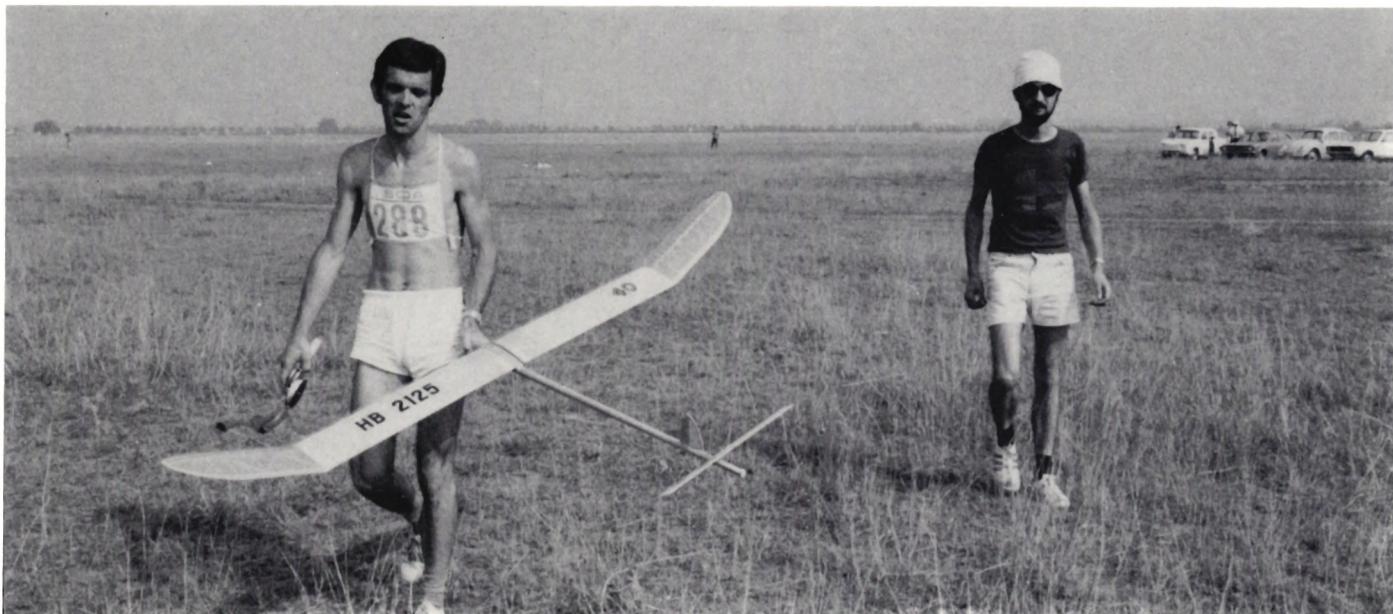 A black and white photograph of two men in a grassy field. The man on the left, Toni Bucher, is wearing a white athletic singlet with the number '289' and white shorts. He is carrying a large, light-colored glider with the text 'HB 2125' on its fuselage. The man on the right is wearing a dark t-shirt, light shorts, and a white cap, and is walking towards the camera. In the background, there are several parked vehicles and a distant horizon line.