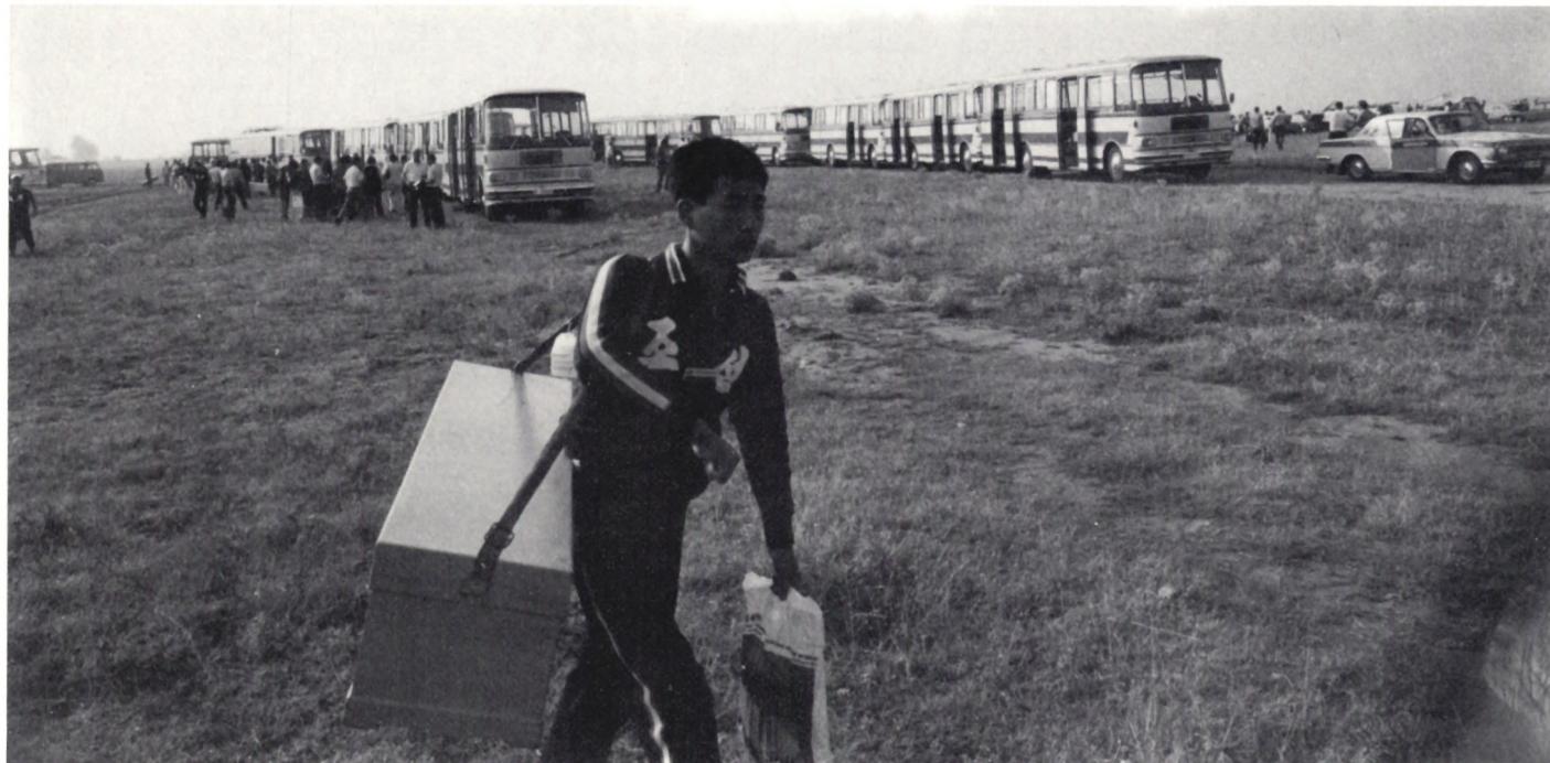 A black and white photograph showing a long line of buses parked on a grassy field. In the foreground, a man in a dark tracksuit is walking, carrying a large, light-colored rectangular box. Other people are visible in the background near the buses.