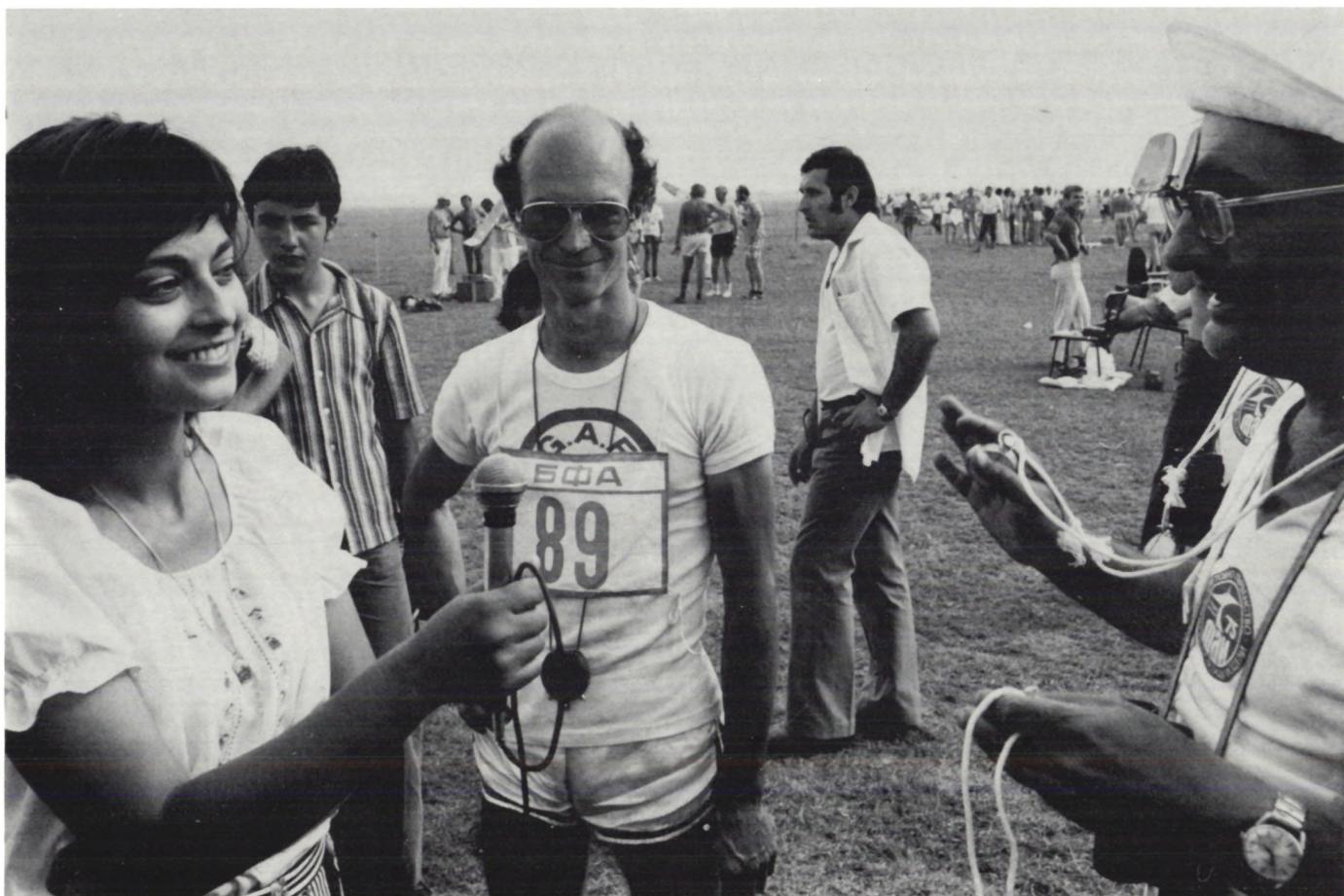 A black and white photograph of a woman interviewing Urs Schaller. The woman is on the left, smiling and holding a microphone. Urs Schaller is in the center, wearing a white t-shirt with a logo and the number 89. He is also wearing sunglasses and a necklace. To the right, another man is partially visible, also wearing sunglasses and a necklace. The background shows a crowd of spectators in a grassy field.