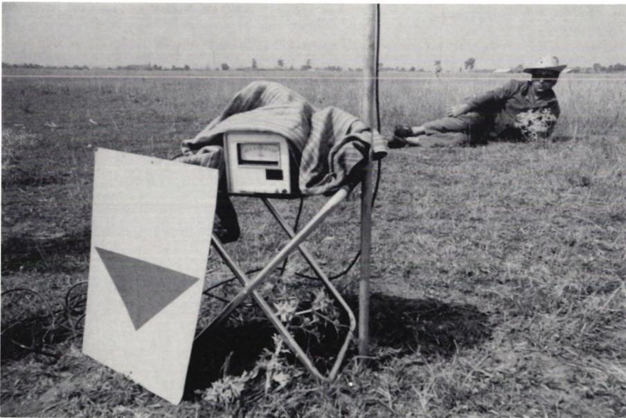 A person lying on the ground in a field, possibly a pilot or crew member, next to a piece of equipment on a tripod.