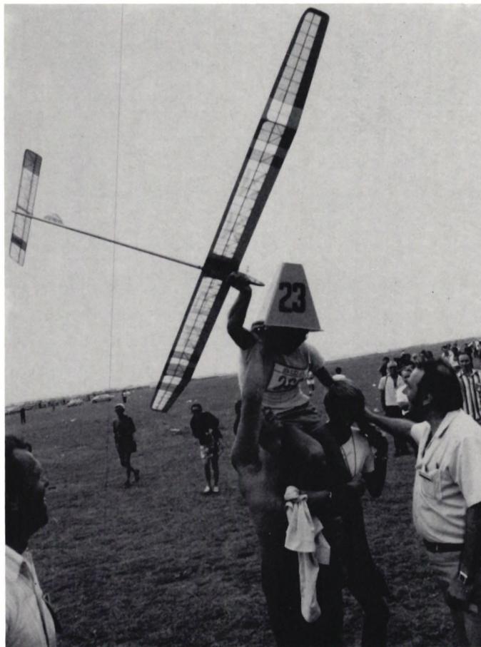 A black and white photograph of a man, Viktor Tchop, being celebrated. He is wearing a white athletic singlet with the number '23' and a white cap. He is being held up on the shoulders of several people. A large, light-colored glider is being held above him. A crowd of spectators is visible in the background, and the sky is clear.