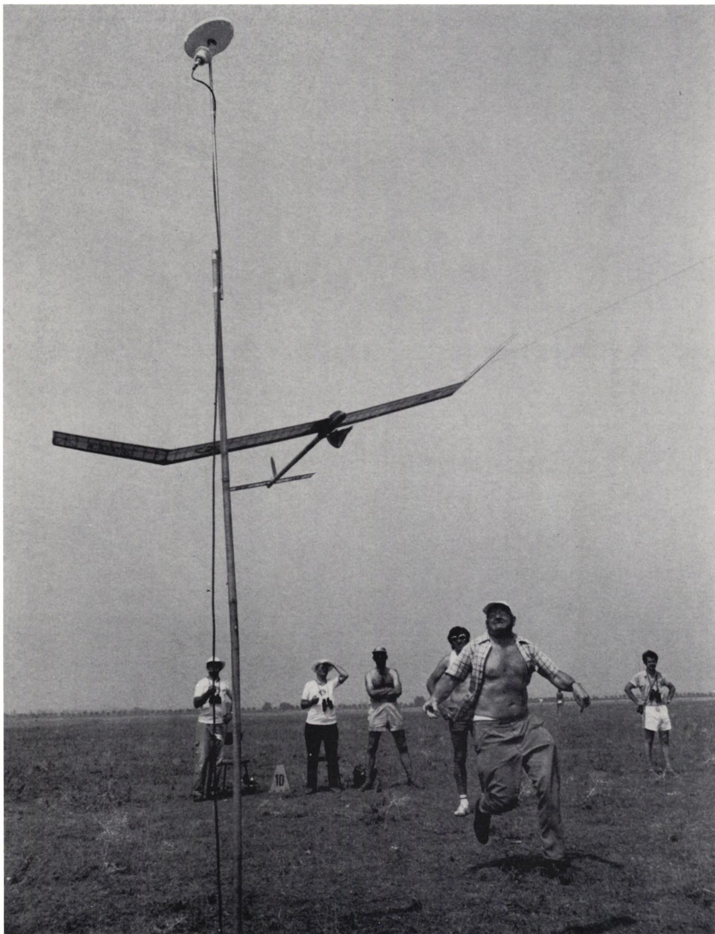 A black and white photograph of a model airplane on a tall pole in a field, with several people watching.