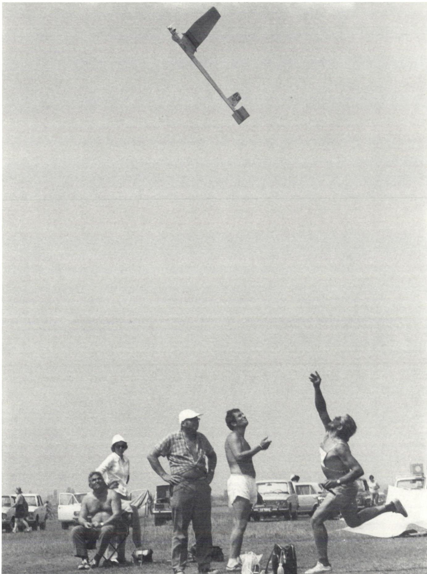 A black and white photograph capturing a moment at a model aircraft competition. In the upper center, a small, high-wing model airplane is captured in mid-flight against a clear sky. Below, on a grassy field, a group of people are gathered. On the right, a man in athletic wear is captured in the middle of a long-throw spear throw, his body twisted in motion. To his left, several other men are watching the event; one is standing with hands on hips, another is sitting on a folding chair. In the background, several cars are parked, suggesting a casual, outdoor setting. The overall atmosphere is one of focused activity and competition.
