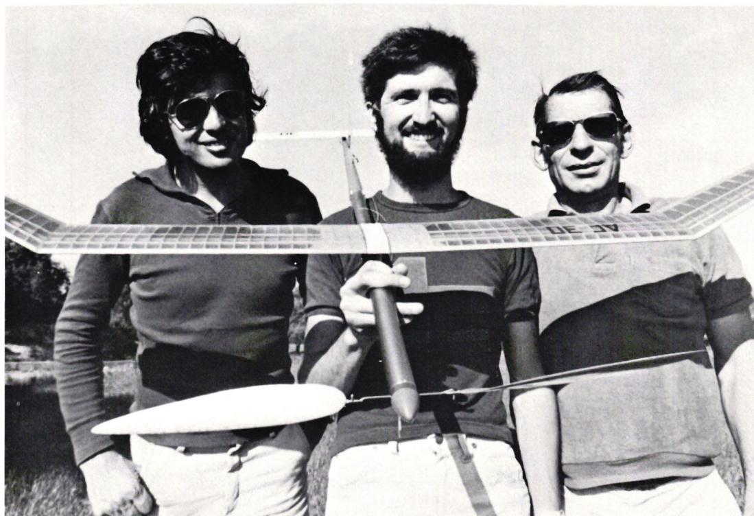 A black and white photograph of three men standing outdoors, each holding a model airplane. From left to right: Siebenmann, Reifler, and Gänsli. They are all smiling and wearing sunglasses. The man in the center, Reifler, is holding a model airplane with a large white wing and a dark body. The other two men are also holding similar model airplanes. The background is a bright, slightly hazy sky.