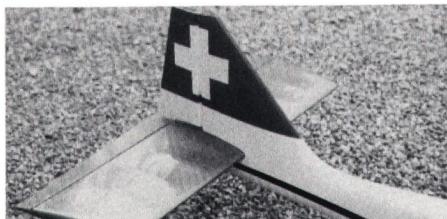Close-up photograph of the tail section of a model aircraft, showing the vertical and horizontal stabilizers with a white cross symbol on a dark background.