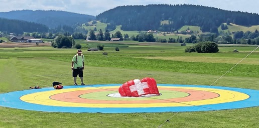 A person standing on a large, circular, multi-colored target mat (yellow, orange, red, blue) in a grassy field. A red and white parachute is visible on the mat. In the background, there are rolling green hills and a small building.