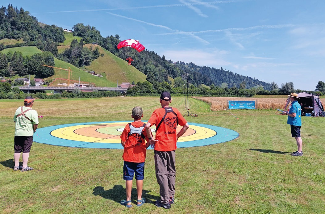 A wide-angle shot of the competition area. In the foreground, three people (two adults and a child) are watching a large, circular, multi-colored target (yellow, orange, red, blue) on a grassy field. In the background, a red and white parachute is visible in the sky, and a small structure or tent is set up on the right side.